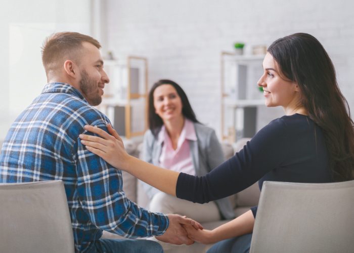 A couple sits facing each other in a therapy session, holding hands. The woman is smiling at her partner, while the man looks happy and relaxed. A lady counselor sits behind them