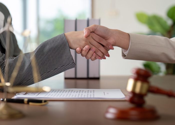 Two lawyers shaking hands over a desk with legal documents, scales of justice, and a gavel, symbolizing agreement and partnership.